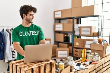 Poster - Young hispanic man wearing volunteer uniform using laptop at charity center