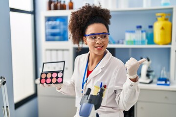 Wall Mural - Young african american woman working at scientist laboratory doing make up pointing thumb up to the side smiling happy with open mouth