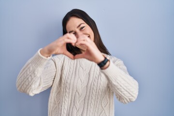 Canvas Print - Young brunette woman standing over blue background smiling in love doing heart symbol shape with hands. romantic concept.