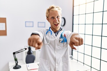 Wall Mural - Young blond man wearing doctor uniform and stethoscope at clinic pointing to you and the camera with fingers, smiling positive and cheerful