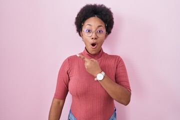 Sticker - Beautiful african woman with curly hair standing over pink background surprised pointing with finger to the side, open mouth amazed expression.