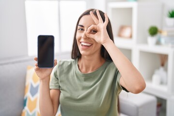 Poster - Young brunette woman holding smartphone showing blank screen smiling happy doing ok sign with hand on eye looking through fingers