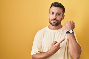 Sticker - Handsome hispanic man standing over yellow background in hurry pointing to watch time, impatience, looking at the camera with relaxed expression