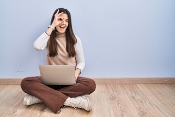 Poster - Young brunette woman working using computer laptop sitting on the floor doing ok gesture with hand smiling, eye looking through fingers with happy face.