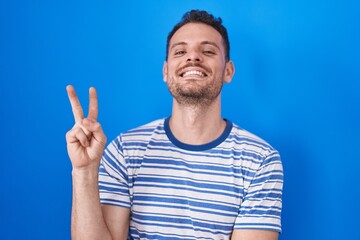 Canvas Print - Young hispanic man standing over blue background smiling with happy face winking at the camera doing victory sign with fingers. number two.