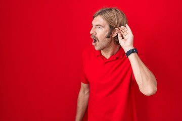 Poster - Caucasian man with mustache standing over red background smiling with hand over ear listening an hearing to rumor or gossip. deafness concept.