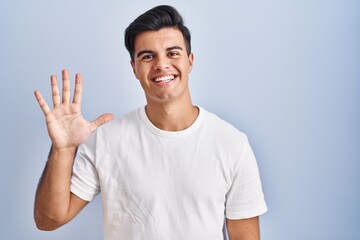 Wall Mural - Hispanic man standing over blue background showing and pointing up with fingers number five while smiling confident and happy.