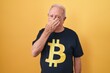 Senior man with grey hair wearing bitcoin t shirt smelling something stinky and disgusting, intolerable smell, holding breath with fingers on nose. bad smell