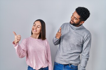 Poster - Young hispanic couple standing together looking proud, smiling doing thumbs up gesture to the side