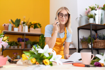 Poster - Young blonde woman florist talking on smartphone reading book at flower shop