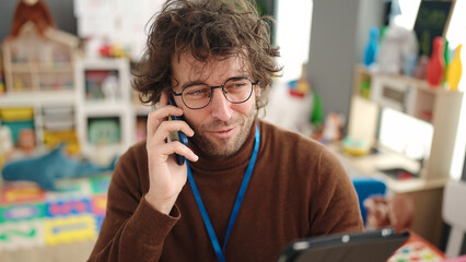 Poster - Young hispanic man preschool teacher using touchpad talking on smartphone at kindergarten