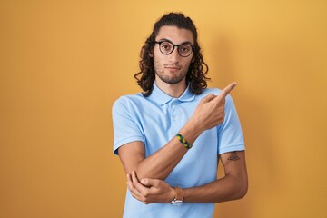 Poster - Young hispanic man standing over yellow background pointing with hand finger to the side showing advertisement, serious and calm face