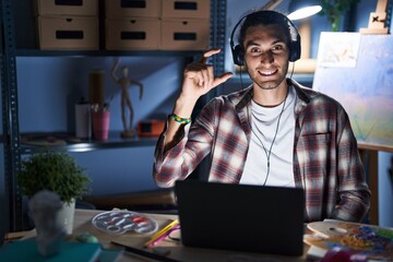 Poster - Young hispanic man sitting at art studio with laptop late at night smiling and confident gesturing with hand doing small size sign with fingers looking and the camera. measure concept.