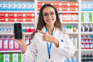 Wall Mural - Young hispanic woman working at pharmacy drugstore showing smartphone screen clueless and confused expression. doubt concept.