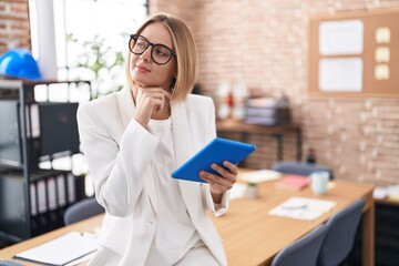 Wall Mural - Young caucasian woman working at the office wearing glasses with hand on chin thinking about question, pensive expression. smiling with thoughtful face. doubt concept.