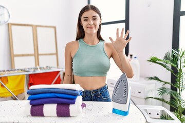 Canvas Print - Young hispanic woman ironing clothes at laundry room showing and pointing up with fingers number five while smiling confident and happy.