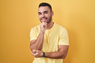 Sticker - Young hispanic man standing over yellow background with hand on chin thinking about question, pensive expression. smiling and thoughtful face. doubt concept.