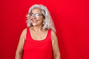 Canvas Print - Middle age woman with grey hair standing over red background looking away to side with smile on face, natural expression. laughing confident.