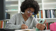© Krakenimages.com - African american woman student using smartphone writing on notebook at library university