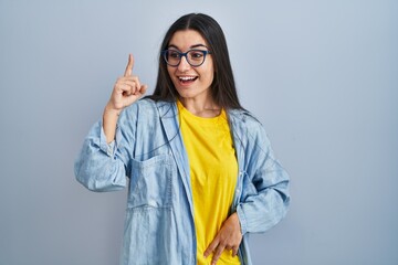 Canvas Print - Young hispanic woman standing over blue background pointing finger up with successful idea. exited and happy. number one.