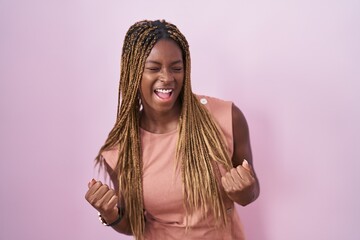 Poster - African american woman with braided hair standing over pink background very happy and excited doing winner gesture with arms raised, smiling and screaming for success. celebration concept.