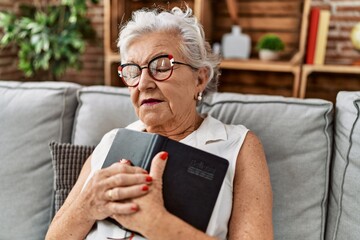 Wall Mural - Senior grey-haired woman hugging bible sitting on sofa at home