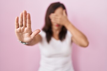 Poster - Middle age brunette woman standing over pink background covering eyes with hands and doing stop gesture with sad and fear expression. embarrassed and negative concept.