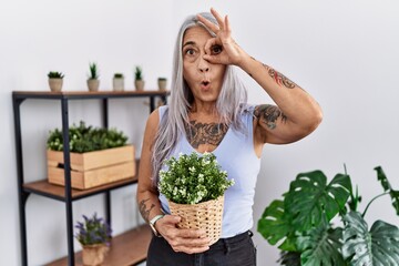 Sticker - Middle age grey-haired woman holding green plant pot at home doing ok gesture shocked with surprised face, eye looking through fingers. unbelieving expression.