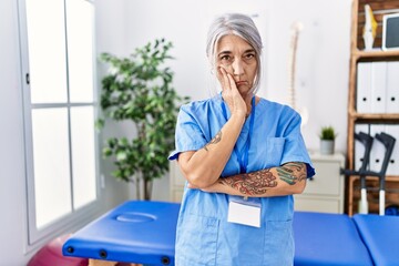 Wall Mural - Middle age grey-haired woman wearing physiotherapist uniform at medical clinic thinking looking tired and bored with depression problems with crossed arms.