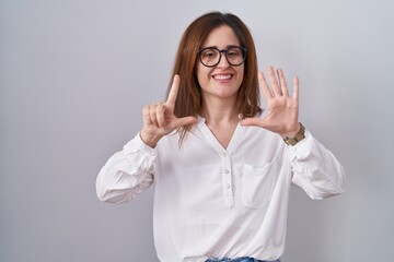 Sticker - Brunette woman standing over white isolated background showing and pointing up with fingers number seven while smiling confident and happy.