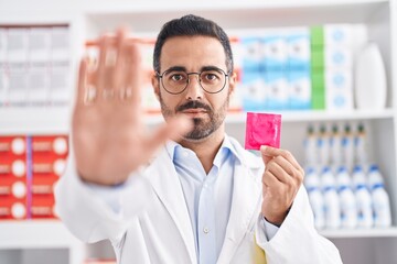 Wall Mural - Hispanic man with beard working at pharmacy drugstore holding condom with open hand doing stop sign with serious and confident expression, defense gesture