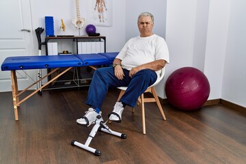 Wall Mural - Senior caucasian man at physiotherapy clinic using pedal exerciser puffing cheeks with funny face. mouth inflated with air, crazy expression.