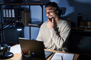 Canvas Print - Young handsome man working using computer laptop at night looking confident at the camera smiling with crossed arms and hand raised on chin. thinking positive.