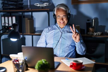 Wall Mural - Hispanic senior man wearing call center agent headset at night smiling with happy face winking at the camera doing victory sign. number two.