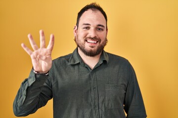 Wall Mural - Plus size hispanic man with beard standing over yellow background showing and pointing up with fingers number four while smiling confident and happy.