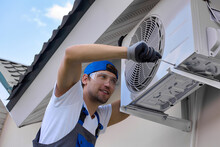 Experienced Craftsman With Smile Tightens Bolts Body Air Conditioner Unit Under Roof Of Building With A Screwdriver. Reliable Installation Of Outdoor Air Conditioning On The Facade Of The Building