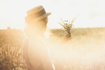 Wall Mural - Close up girl in straw hat with bouquet surrounded by sunlight glare concept photo. Side view photography with meadow on background. High quality picture for wallpaper, travel blog, magazine, article