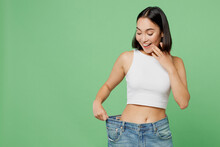 Young Smiling Happy Woman Wears White Clothes Show Loose Pants On Waist After Weightloss Isolated On Plain Pastel Light Green Background. Proper Nutrition Healthy Fast Food Unhealthy Choice Concept.