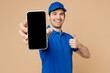 © ViDi Studio - Delivery guy employee man wearing blue cap t-shirt uniform workwear work as dealer courier hold in hand use mobile cell phone with blank screen area show thumb up isolated on plain beige background.