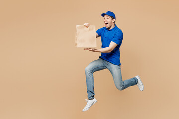 Full body sideways delivery guy employee man wear blue cap t-shirt uniform work as dealer courier jump high hold brown clear blank craft paper takeaway bag mock up isolated on plain beige background.