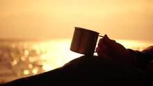 Travel Stainless Steel Mug In The Hands Of A Woman. Small Waves On Golden Warm Water Surface With Bokeh Lights From Sun. A Concept Of The World Of Beauty, Nature And Outdoor Travel. Close Up