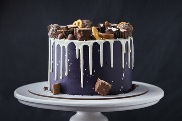 Canvas Print - Birthday cake with dark blue cream cheese frosting with white chocolate drips on the black background. Homemade cake decorated with chocolate sweets and cookies on the white stand