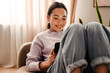 © Drobot Dean - Cheerful asian woman using digital tablet while sitting on sofa in living room