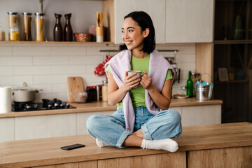 Wall Mural - Cheerful young asian woman drinking coffee while sitting in cozy kitchen