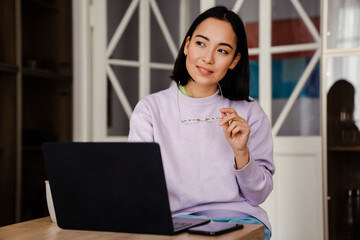 Poster - Pensive asian woman working on laptop while sitting at table at home