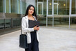 © Clàudia Ayuso - Close up portrait of a young executive businesswoman in urban outdoors holding a folder. Smiling empowered woman dressed smart casual outside the office work site. Horizontal with copy space.
