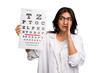 Young indian oculist woman holding an eye chart paper cut out isolated is saying a secret hot braking news and looking aside