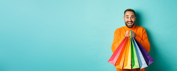 Wall Mural - Excited adult man holding shopping bags and smiling, going to mall, standing over turquoise background
