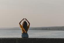 The Young Sexy Girl Sitting With Her Back At The Italian Lake Beach In Yellow T-shirt, Denim Shorts Showing The Heart Symbol With Hands