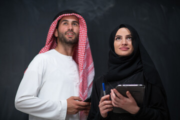 Wall Mural - Young muslim business couple arabian man with woman in fashionable hijab dress using mobile phone and tablet computer in front of black chalkboard representing modern islam fashion technology 